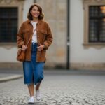Woman wearing brown corduroy jacket with blue denim midi skirt and white sneakers