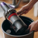 1. Person emptying Dyson stick vacuum dustbin over trash can during routine maintenance cleaning session