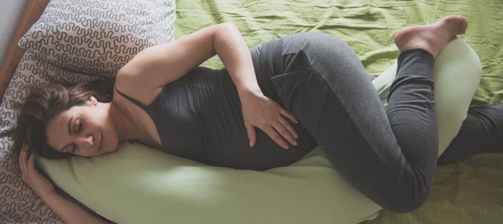 A pregnant woman side-sleeping with a U-shaped pregnancy pillow cradling her belly and back for pain relief.