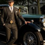 Well-dressed gentleman in 1920s three-piece suit with fedora hat and pocket watch standing beside luxury automobile