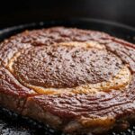 Close-up of seasoned ribeye steak sizzling in hot cast iron skillet with oil glistening on surface