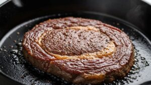 Close-up of seasoned ribeye steak sizzling in hot cast iron skillet with oil glistening on surface