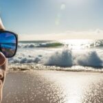 A person wearing polarized anti-glare sunglasses on a sunny beach with ocean waves reflecting bright sunlight in the background