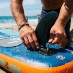 Hands tightening a loose center fin on a paddleboard on a beach.