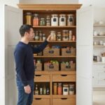 A man reaching for a jar inside a fully organized tall wooden kitchen pantry cabinet.