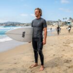 A male surfer on a sunlit beach wearing dark surf leggings with a phone pocket.