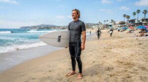 A male surfer on a sunlit beach wearing dark surf leggings with a phone pocket.